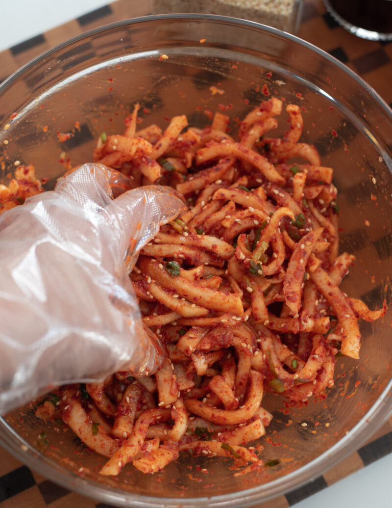 A hand is tossing radish with chili seasoning to make quick radish kimchi in a bowl.