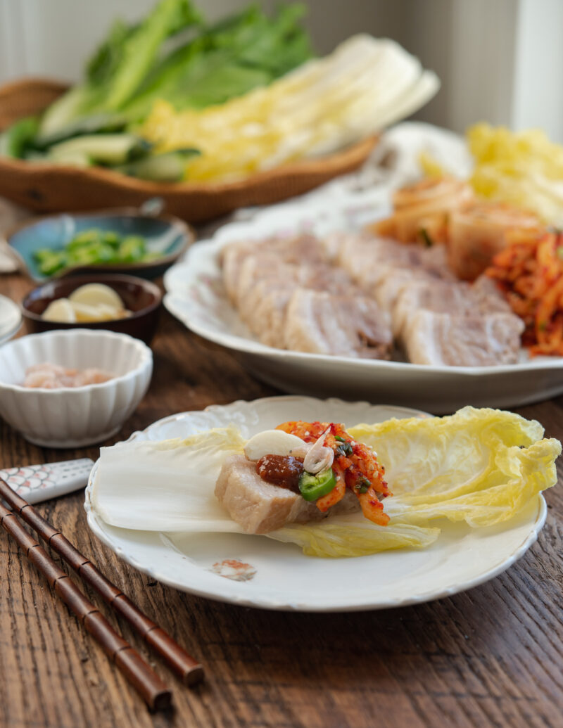 A piece of bossam, radish kimchi, garlic piece are place on a yellow cabbage leaf in a plate.