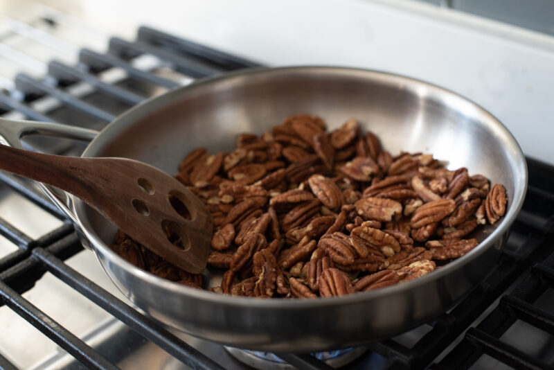 Whole pecan pieces are being toasted in a skillet with a wooden spatula.