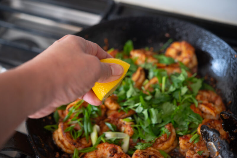 A hand is squeezing out a half lemon to chili shrimp tossed with cilantro.