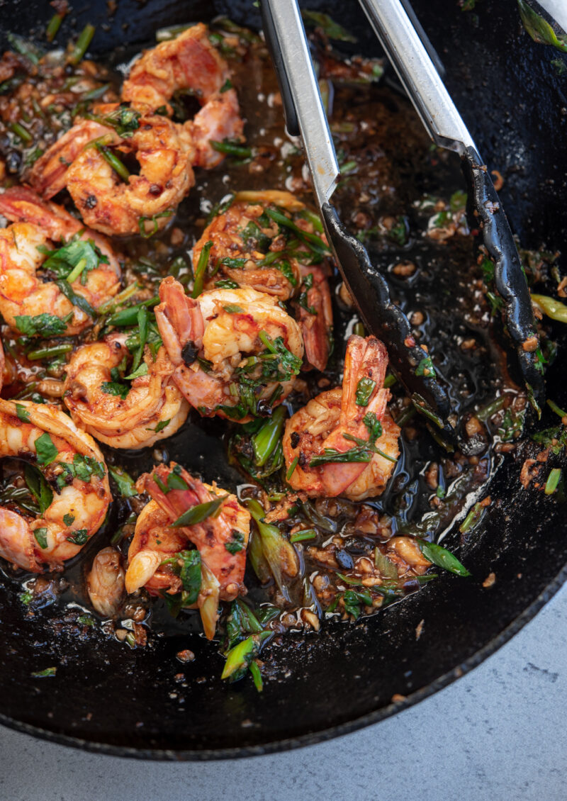 Chili crisp shrimp tossed in cilantro is in the skillet with kitchen tongs.