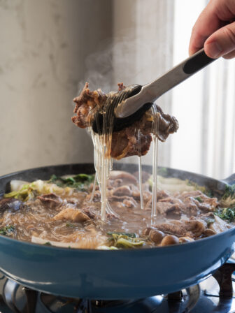 Kitchen tongs are picking up a bulgogi piece with noodles from the simmering hot pot on the portable burner.