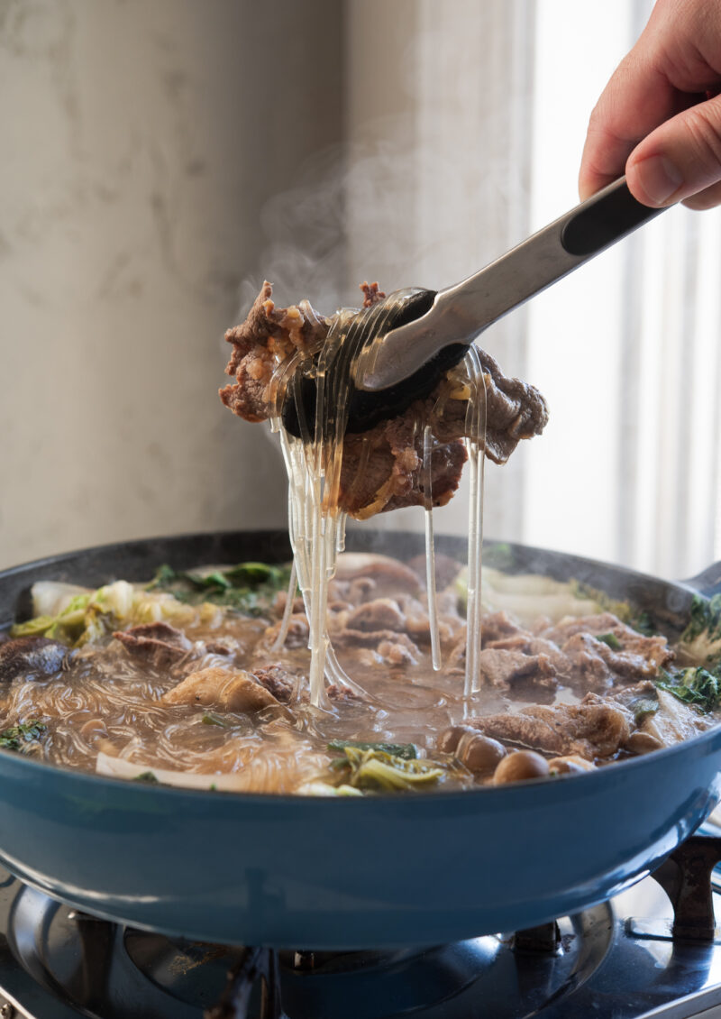 Kitchen tongs are picking up a bulgogi piece with noodles from the simmering hot pot on the portable burner.