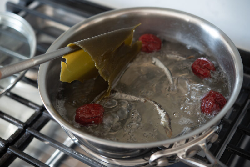 Kithcen tongs are picking up a piece of sea kelp from the stock made with dried anchovies and jujube.