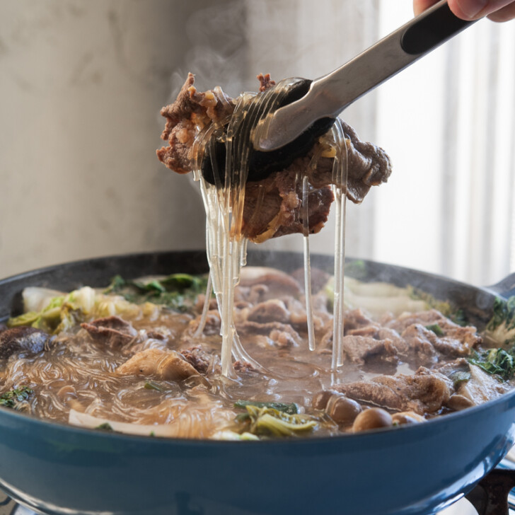 Kitchen tongs are picking up a bulgogi piece with noodles from the simmering hot pot on the portable burner.