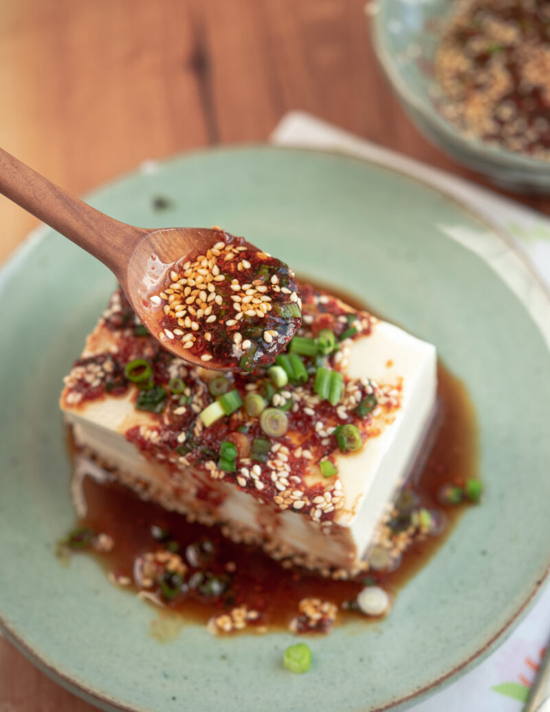 A spoonful of chili soy sauce is drizzling over soft tofu on a green plate.