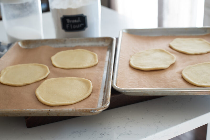 6 flat round doughs are distributed into two baking pans lined with a parchment paper.