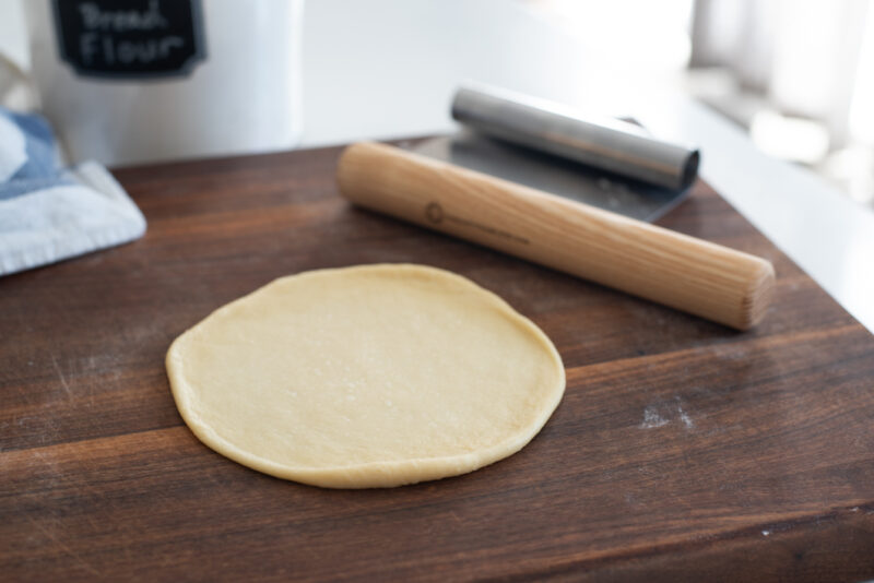 A piece of dough is rolled into thin circle with a mini wooden pin.