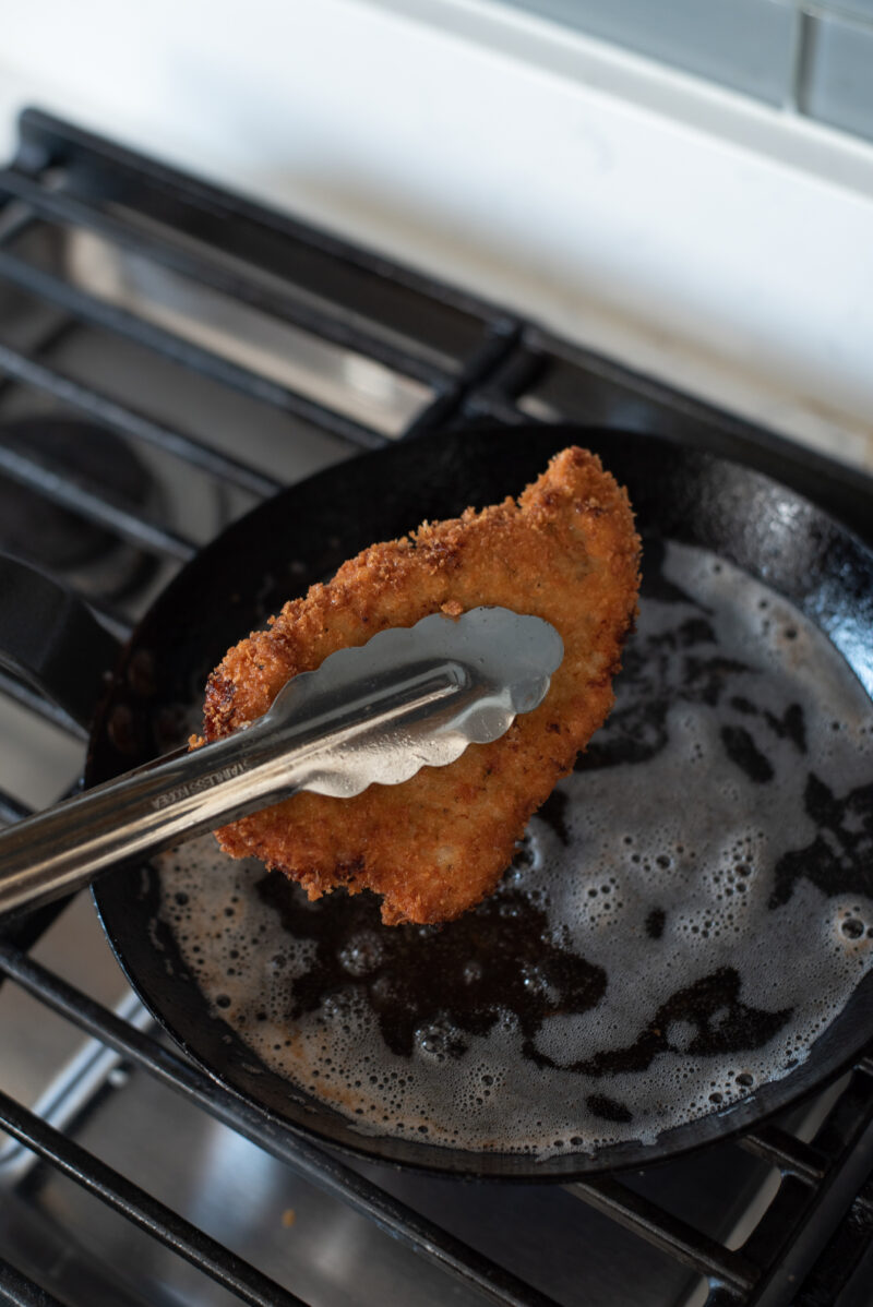 Kitchen tongs are picking up deep fried crispy golden brown tonkatsu piece from the oil in a pan.