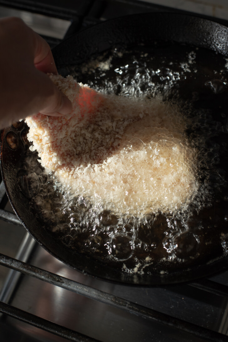 Breaded pork cutlets being lowered into hot oil in a skillet