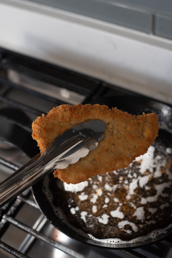 Tongs lifting a golden fried pork cutlet from hot oil
