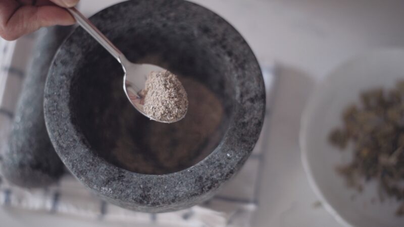 A spoonful of finely grounded cardamom powder is showing over a mortar.