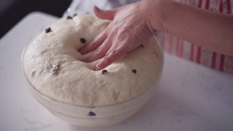 Risen dough in a bowl is punched by a hand to deflate.