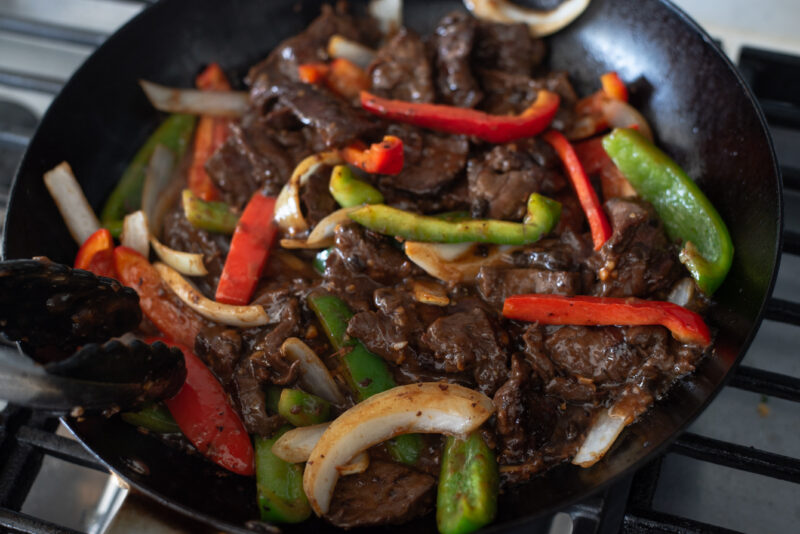 Browned beef strips added to onion and pepper mixture in a skillet.