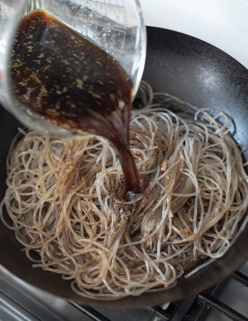 Japchae sauce is poured on to Kroean glass noodles in a pan.