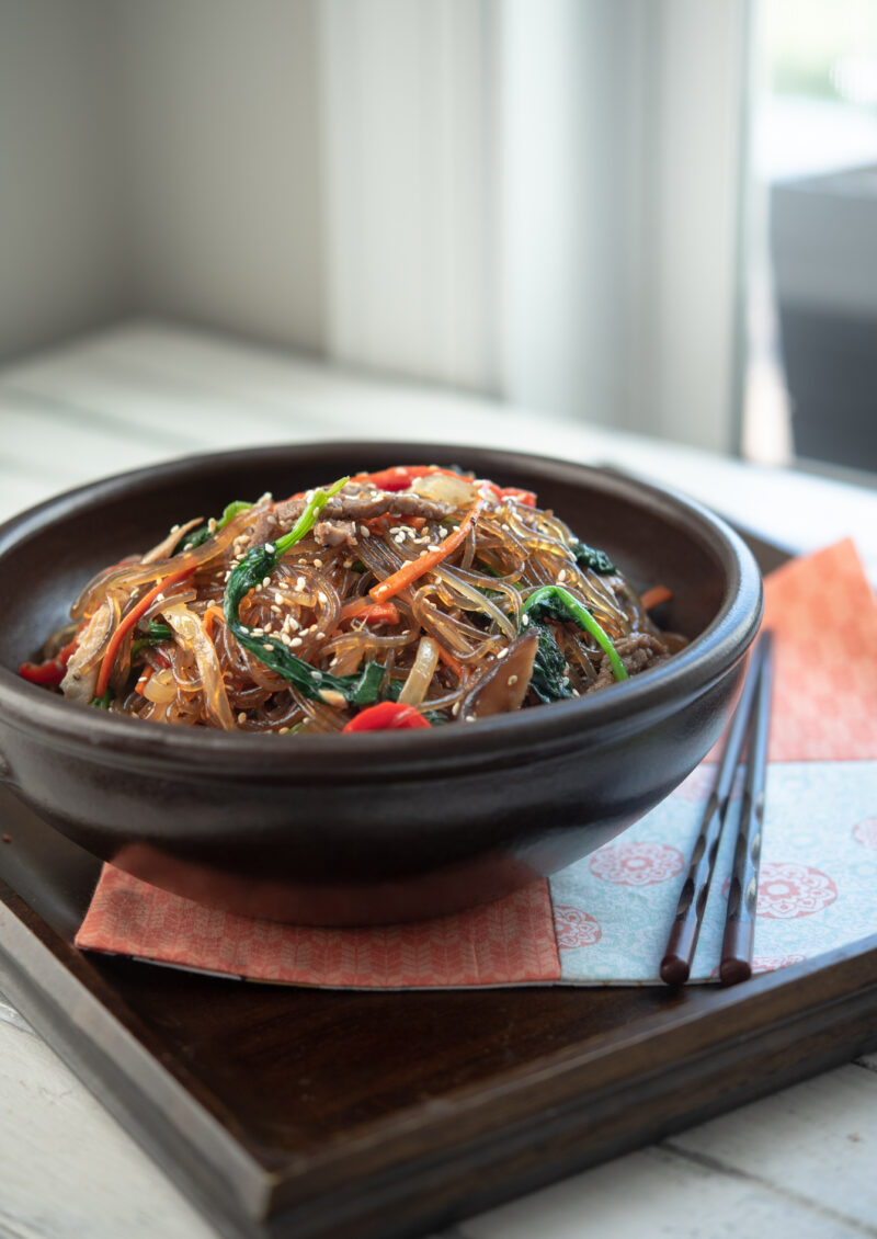 Korean japchae noodles with various vegetables are served in a bowl and chopsticks on a tray.