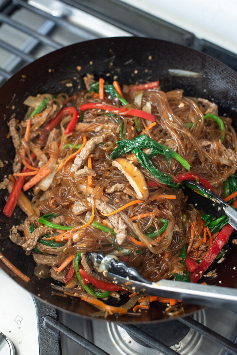 Kitchen tongs are used to mix japchae noodles with vegetables in a skillet.