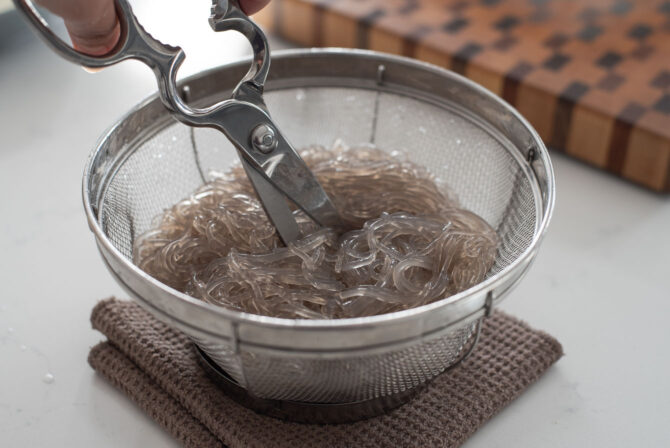 Cooked glass noodles are drained and cut with a scissor