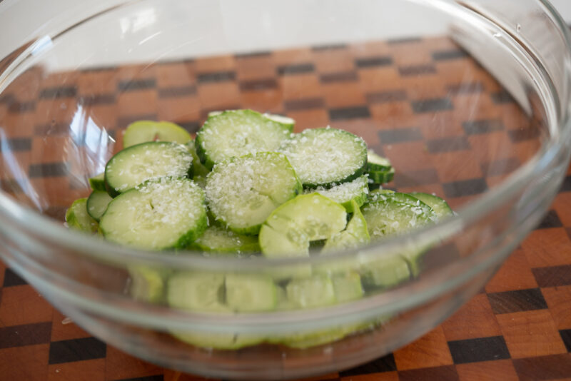A small amount of salt is sprinkled over cucumber slices in a mixing bowl.