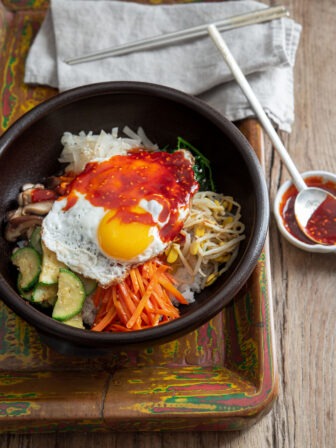 A bowl of Korean bibimbap is on a wooden tray with gochujang sauce on the side