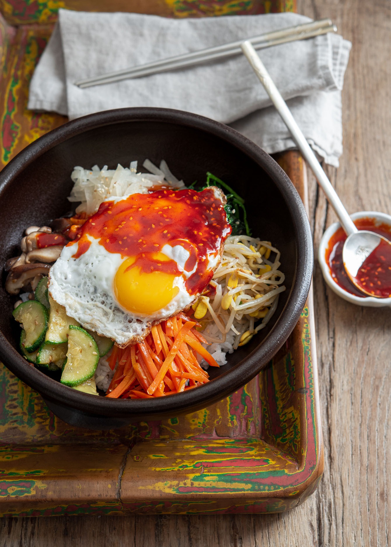 A bowl of Korean bibimbap on a wooden tray served with gochujang sauce on the side.