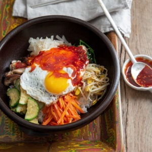 A bowl of Korean bibimbap is on a wooden tray with gochujang sauce on the side
