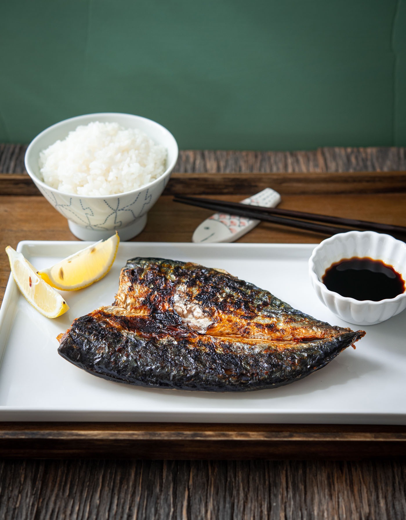 Pan-grilled mackerel fish is on a plate with soy sauce and lemon wedges, and served with rice