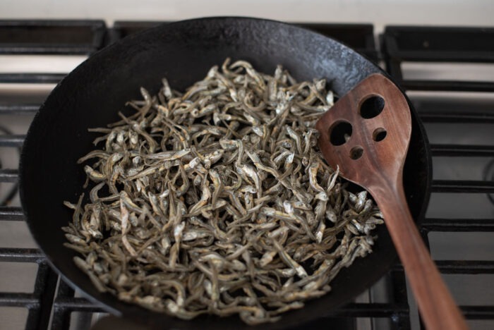 Dried anchovies are toasting in a skillet with a spatula