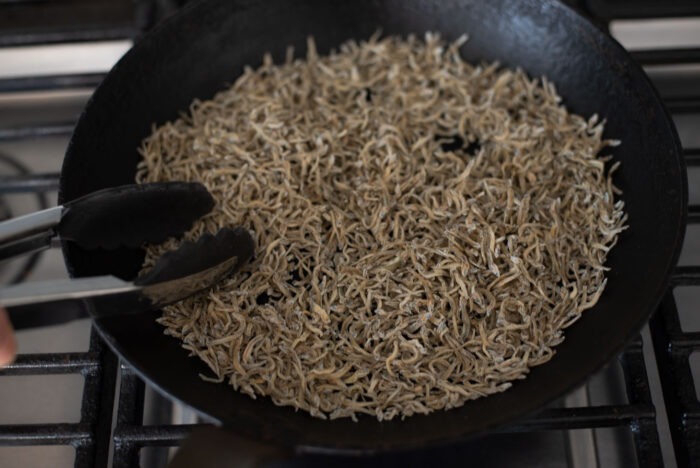 Tiny dried anchovies are toasting in a skillet with a spatula