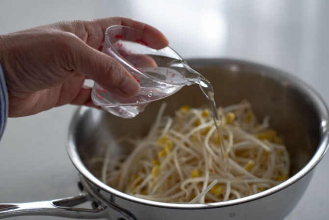 A little cup of water is pouring over soybean sprouts in a pan.