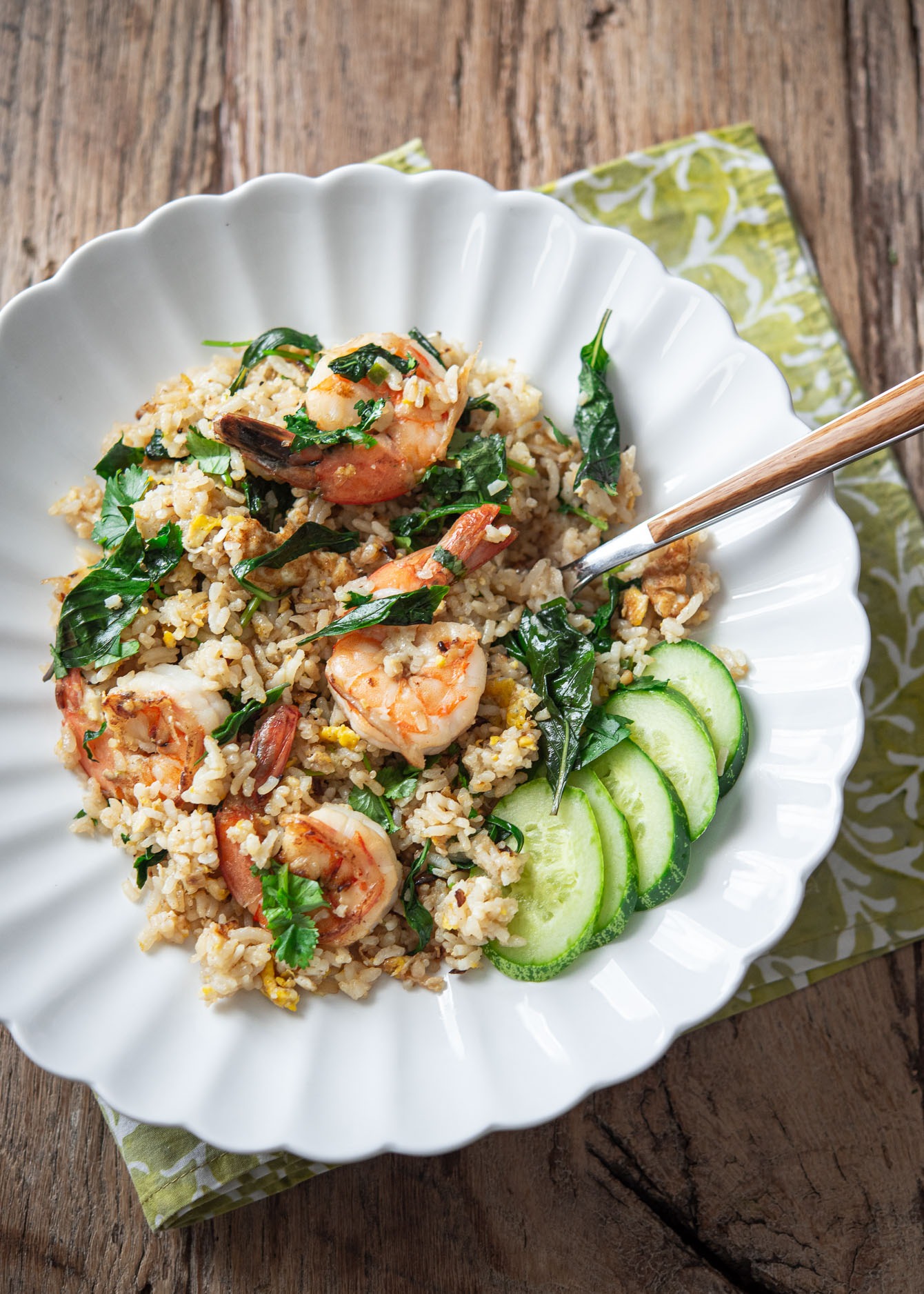 A white plate of Thai basil fried rice and shrimp is served with cucumber slices.