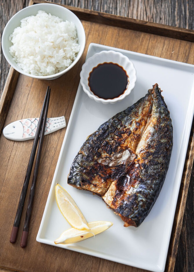 Grilled mackerel fish fillets are on a plate with soy sauce and lemon wedges and served with rice.