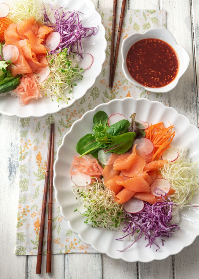 Bowls of bibimbap are arranged with smoked salmon, salad green, and sprouts and served with gochujang sauce