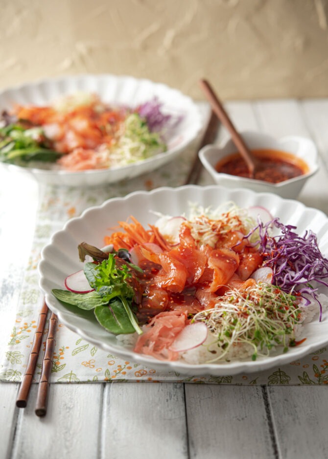 Two white bowls of bibimbap includes smoked salmon and fresh salad vegetables and sprouts