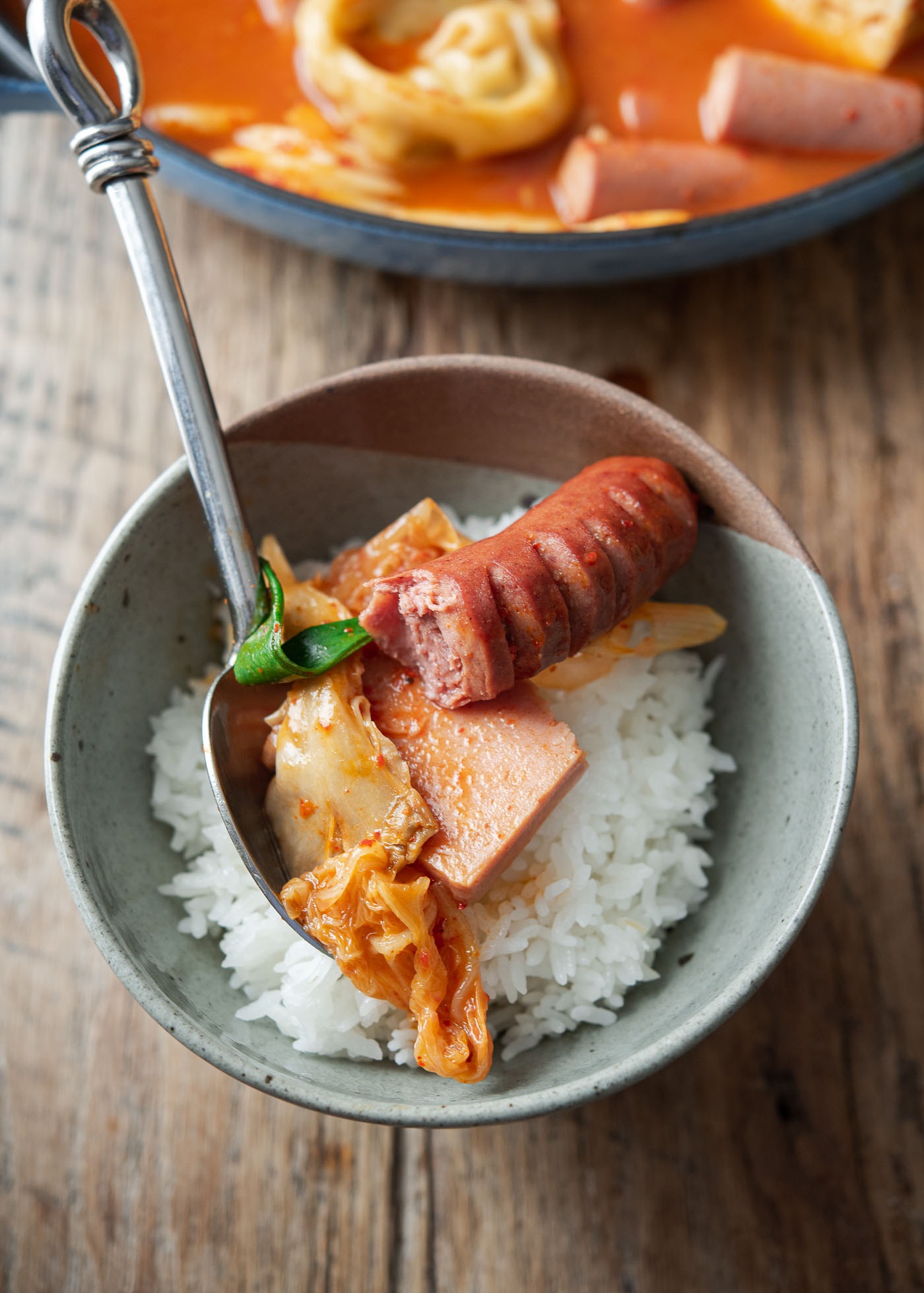 A bowl of rice is topped kimchi budae jjigae Spam, hot dog and kimchi with a spoon.