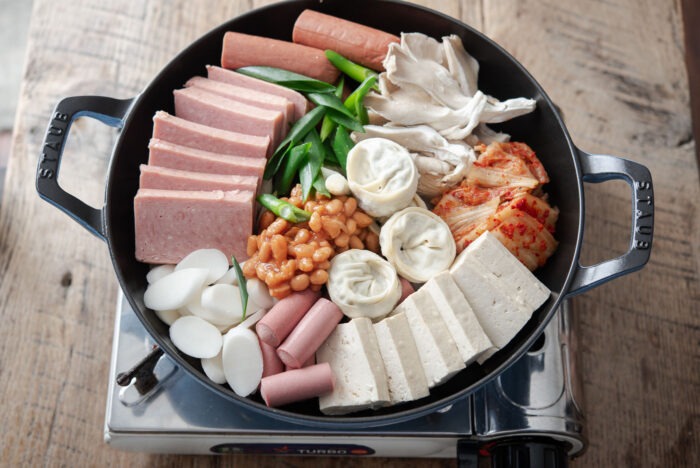 A large pan filled with canned meats, tofu, kimchi and other ingredients are placed on a portable burner.