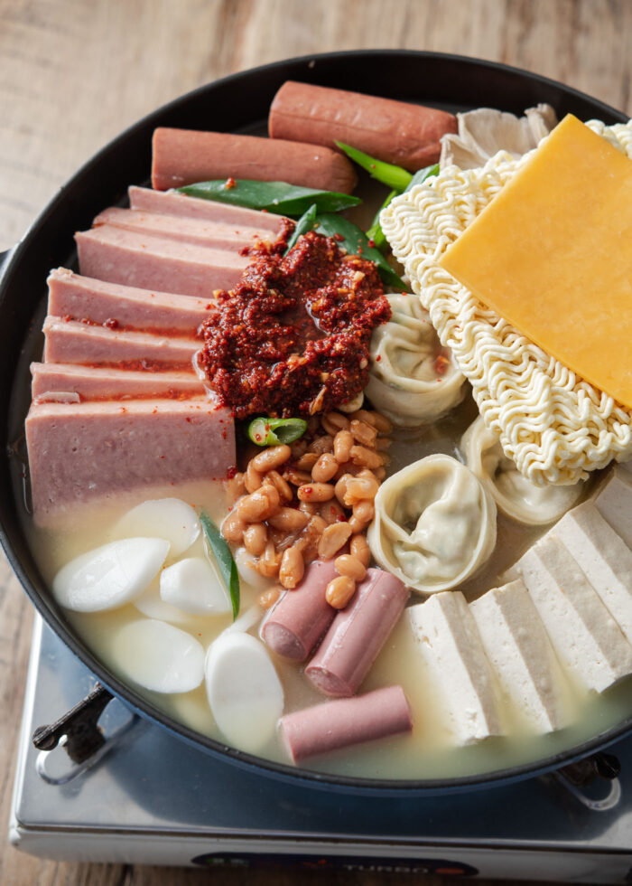 Stock is poured on the budae jjigae pot filled with canned meats, rice cakes, ramen noodles and cheese.