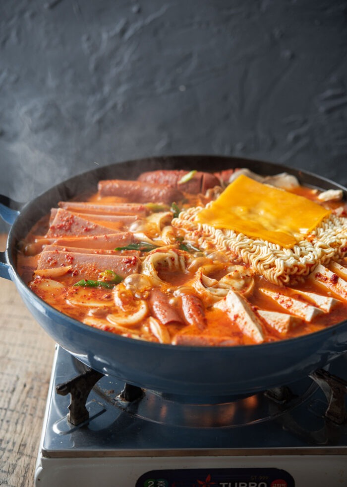 A pot filled with budae jjigae ingredients and ramen noodles are simmering over a portable burner.