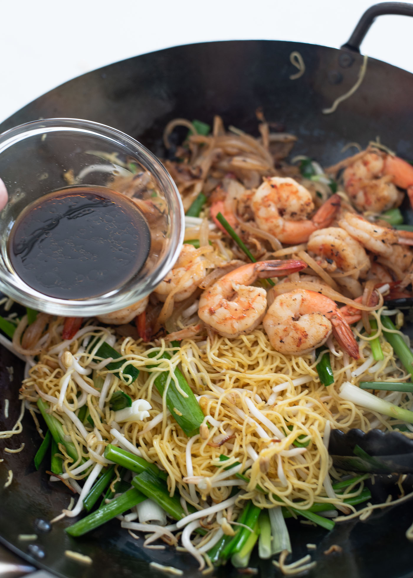 Pan-fried noodle sauce being poured on Hong Kong noodles, shrimp and vegetables in a wok.