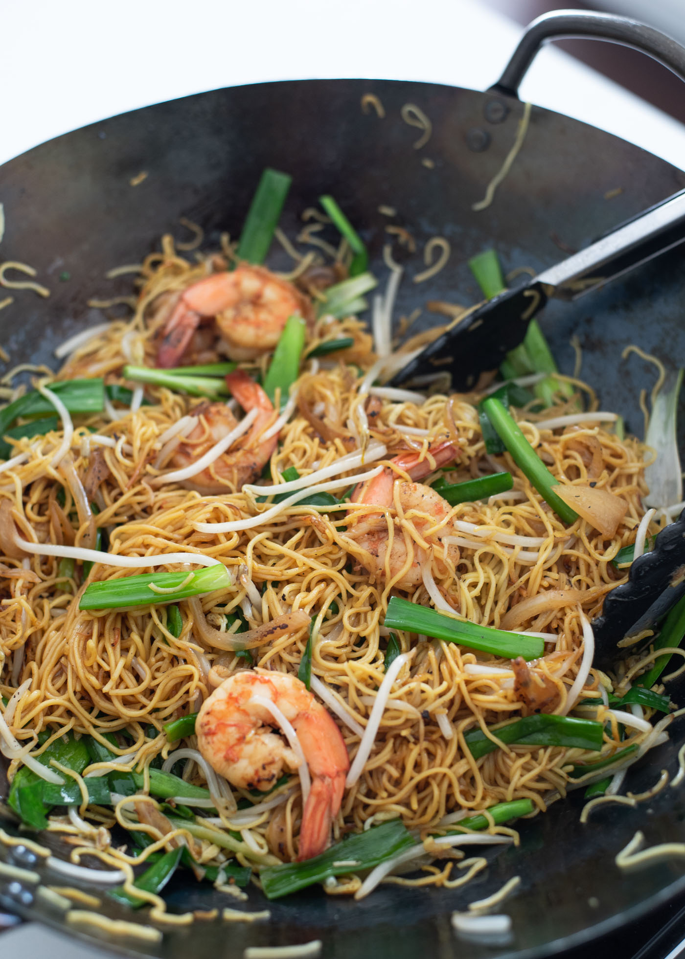 Pan fried egg noodles are tossed together with green onion and shrimp with kitchen tongs in a wok.