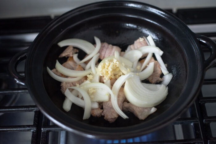 Sliced onion and minced garlic are added to cooked pork belly slices in a pot