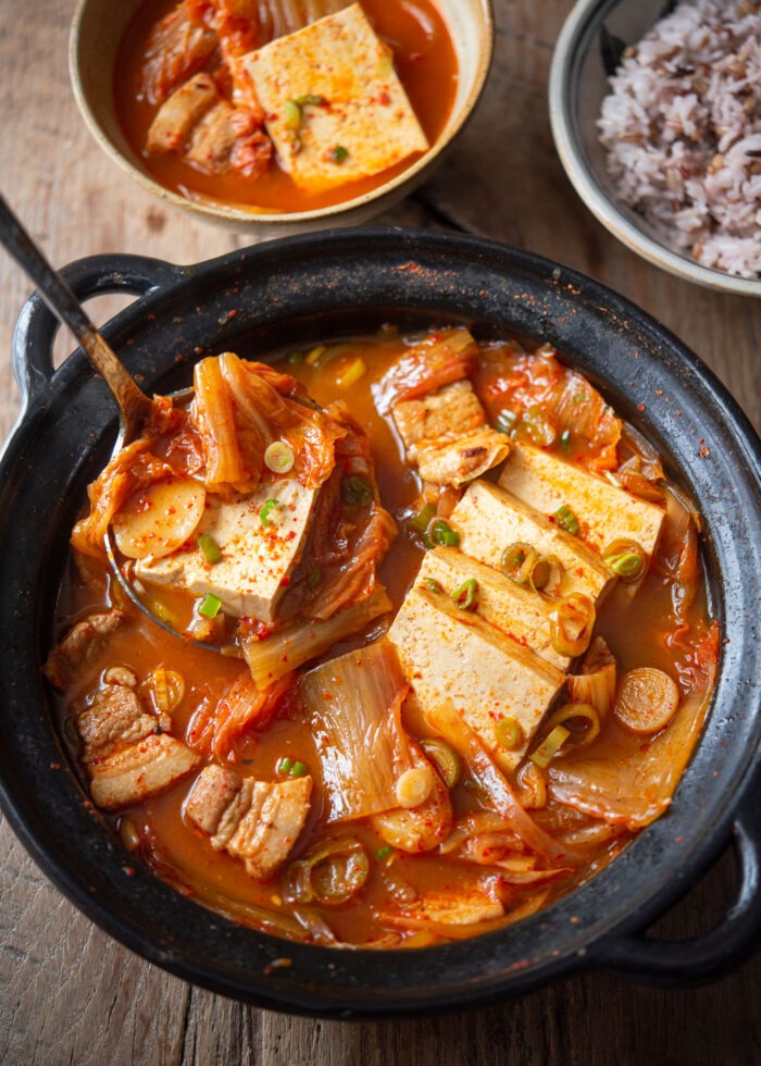 A ladle of kimchi stew is showing slice of tofu, kimchi, and pork from the pot.
