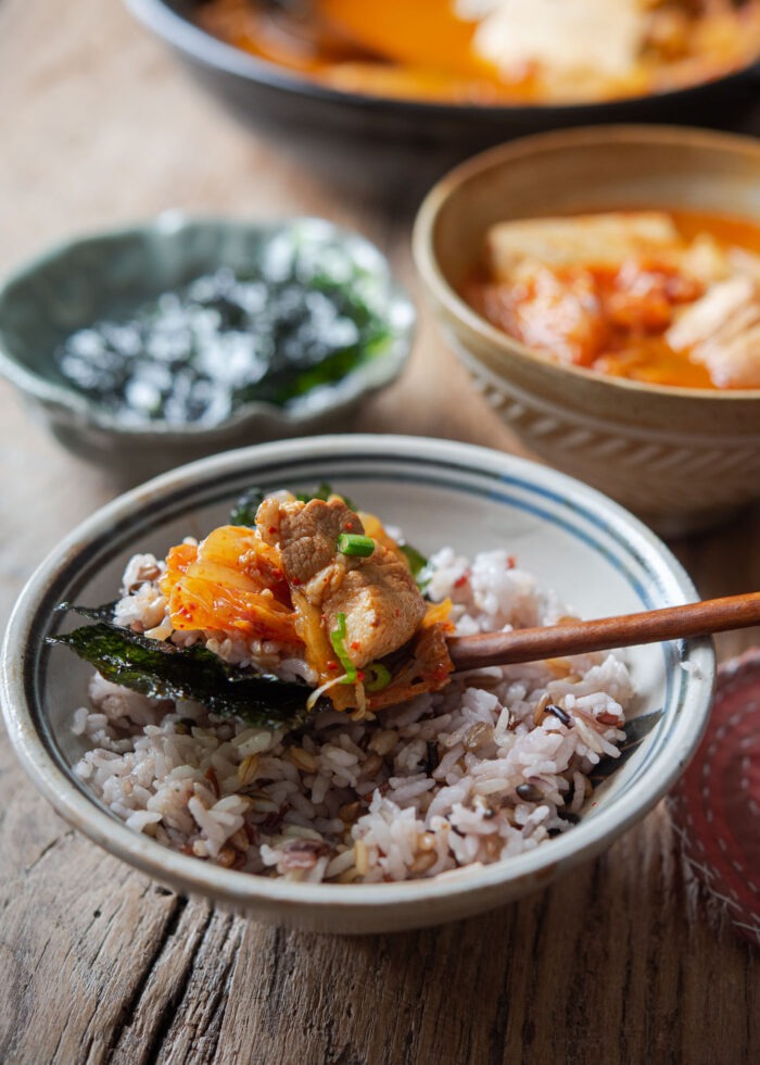 A spoonful of rice is layered with dried seaweed, kimchi stew with pork belly slice on top of rice bowl.