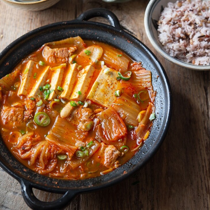 A pot of kimchi jjigae made with pork and tofu is served with rice and roasted seaweed.