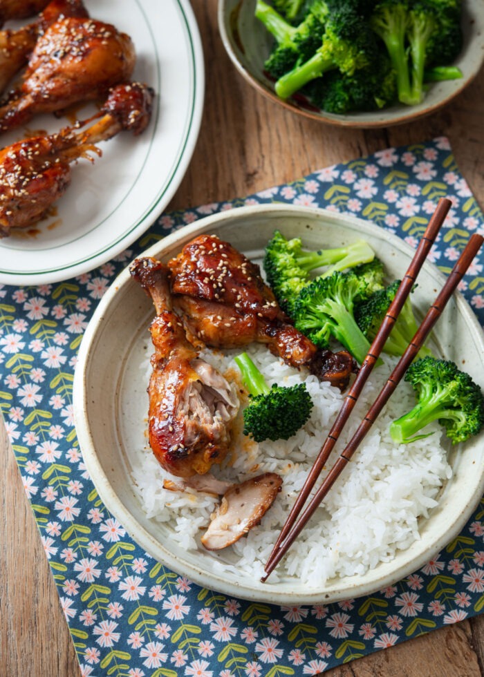Two teriyaki drumsticks are served with rice and steamed broccoli on a plate with chopsticks