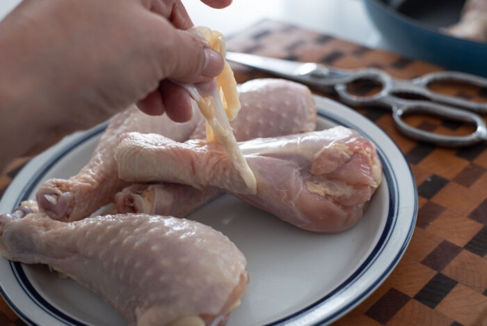 A hand is pulling off the skin removed by kitchen scissors from the chicken drumsticks 