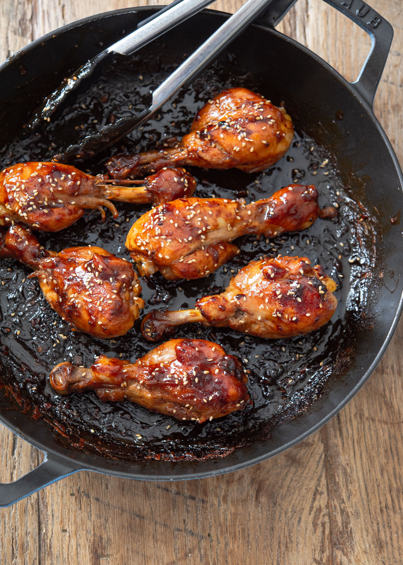 Teriyaki sauce glazed chicken drumsticks are garnished with sesame seeds in a pan with kitchen tongs