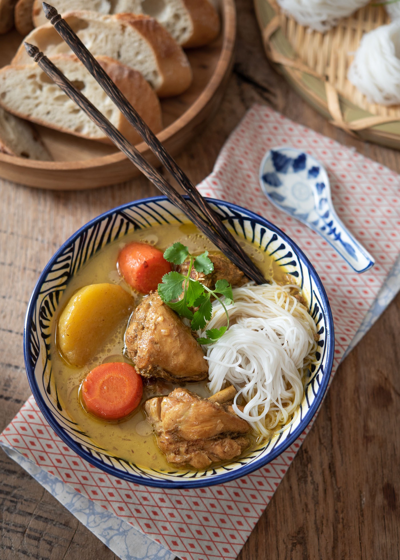 Rice noodles are added in a bowl of chicken, potato, carrot curry and paired with spoon, chopsticks, and baguette slices. 
