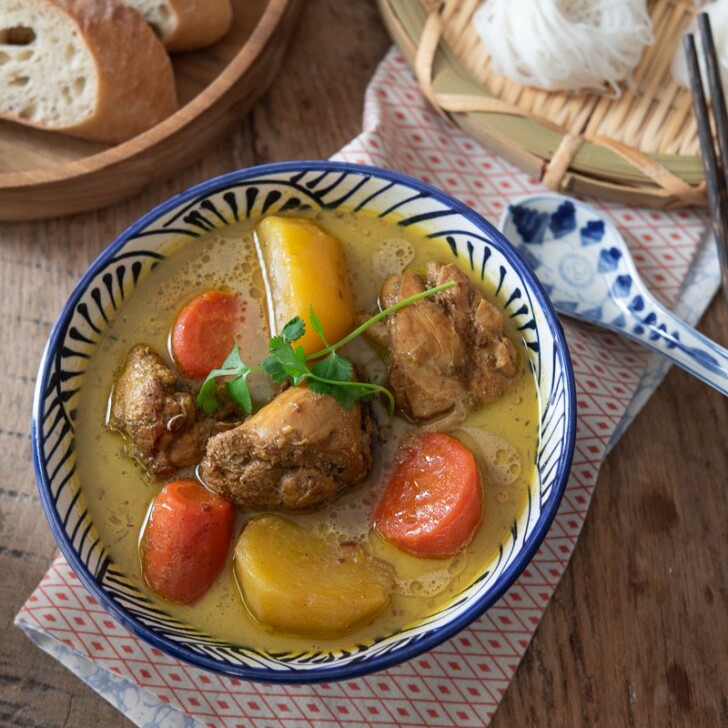 A bowl of chicken curry with potato and carrot are placed on a napkin and paired with baguette slices and rice noodle nests on the side.