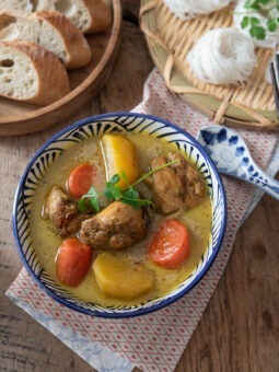 A bowl of chicken curry with potato and carrot are placed on a napkin and paired with baguette slices and rice noodle nests on the side.